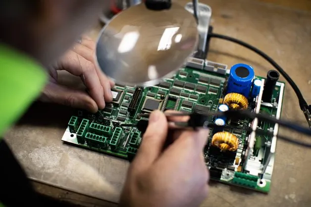 Close-up of hands working on a circuit board under a magnifying lamp